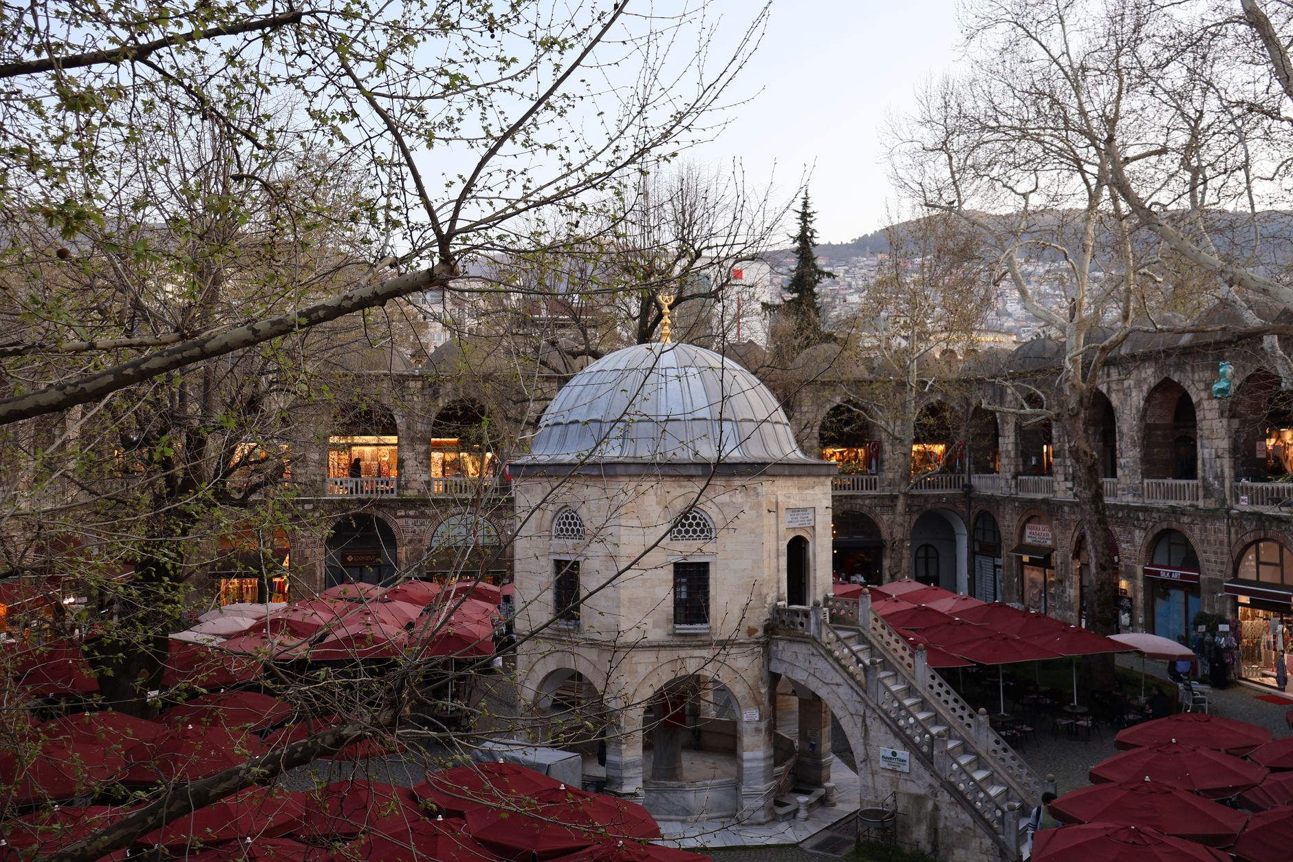A view of the courtyard at Koza Han in Bursa, featuring a dome and red umbrellas among the trees.