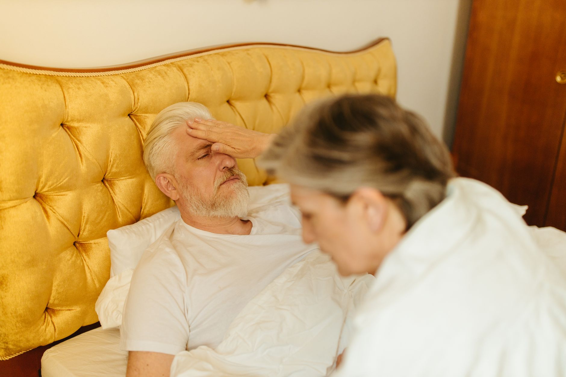 An elderly man resting in bed after surgery, cared for by a woman.