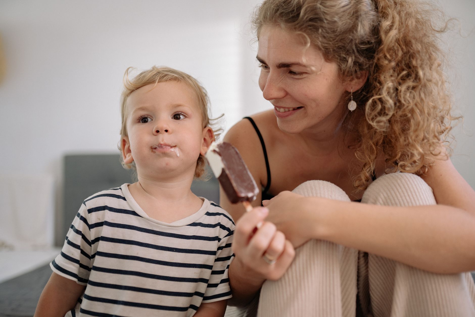 A mother smiles at her child eating ice cream in a calm, cozy room.