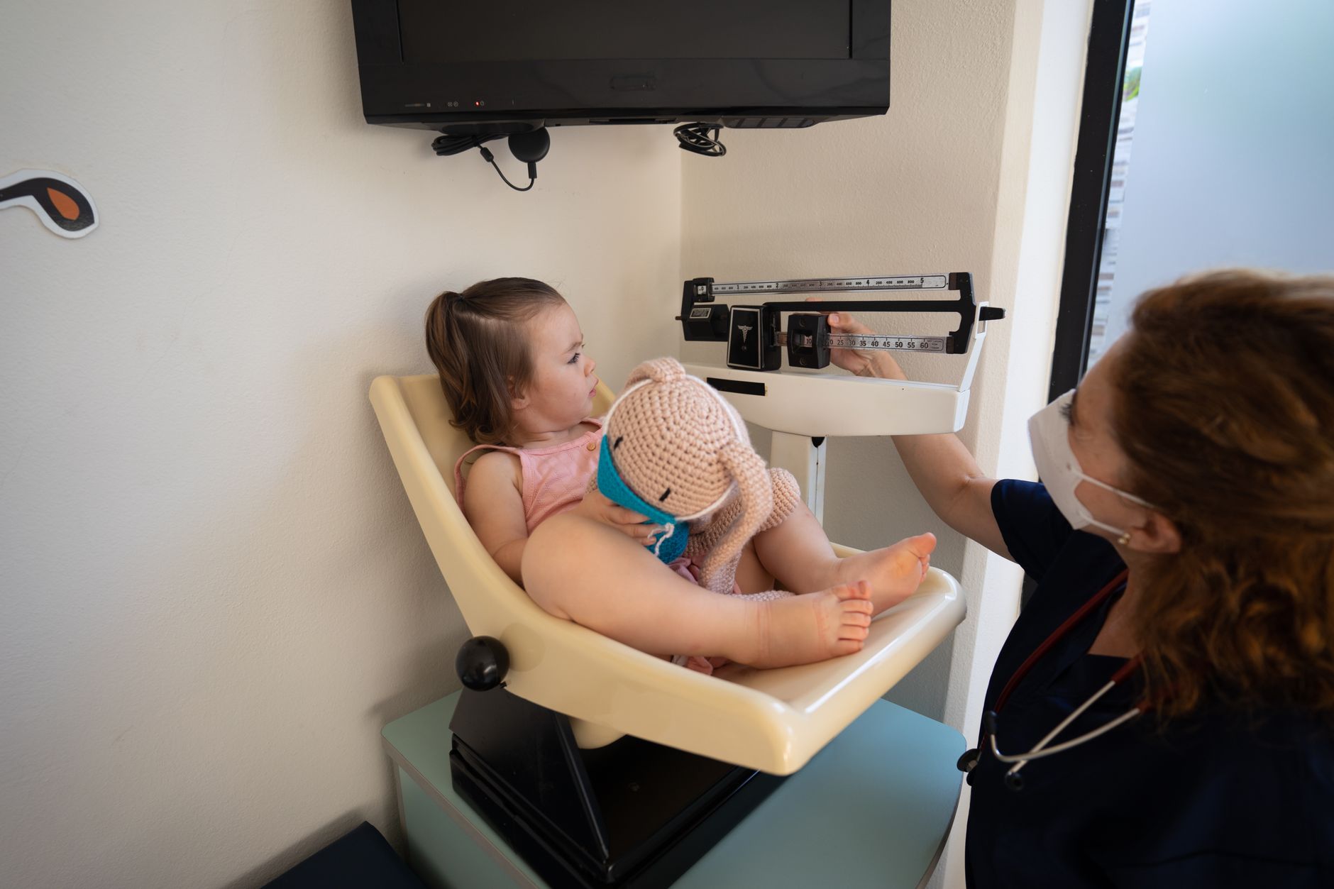 A young child sitting on a scale at a clinic, holding a stuffed toy, with a nurse checking the measurement.