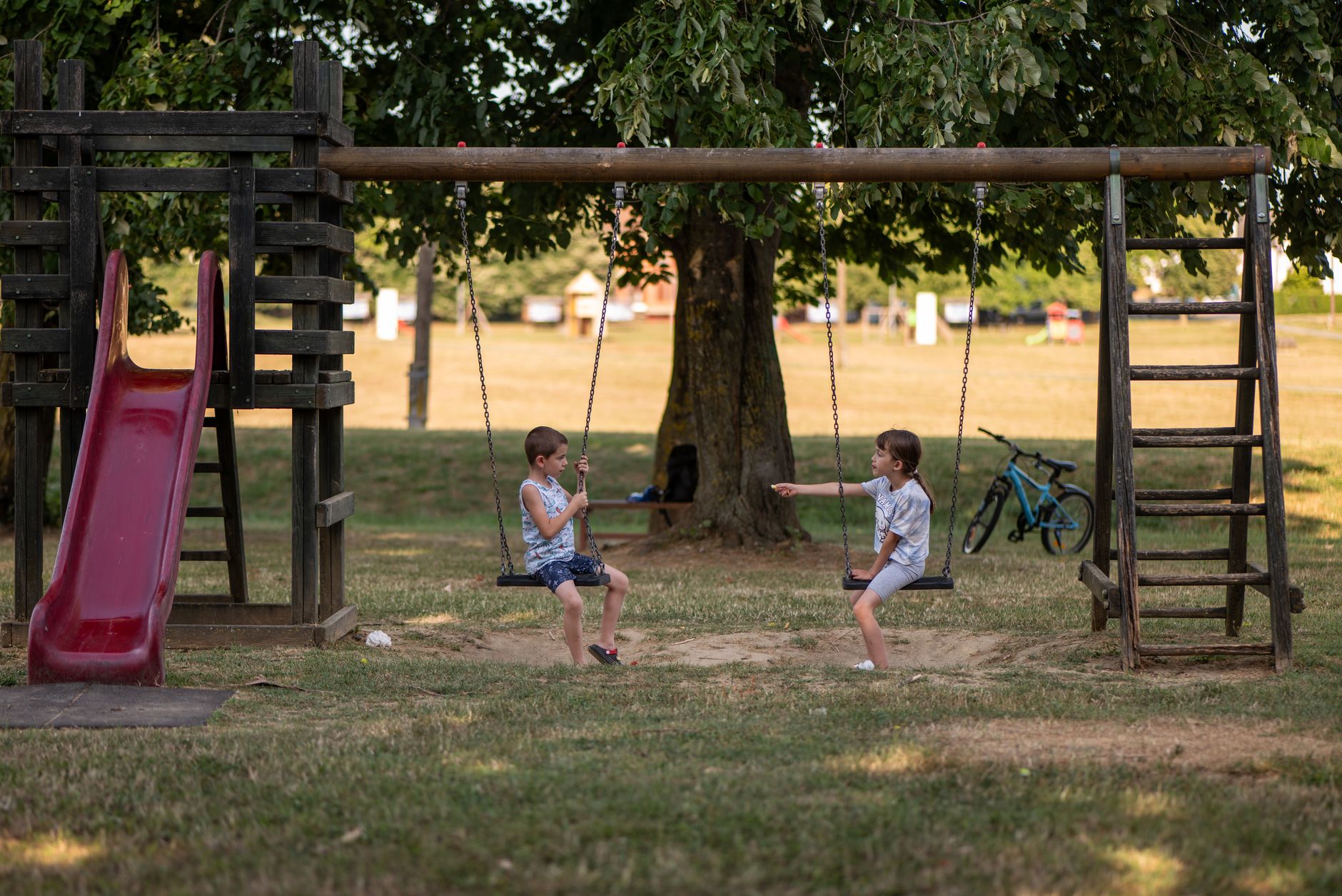 Two children playing on swings in a park, with a slide in the background.