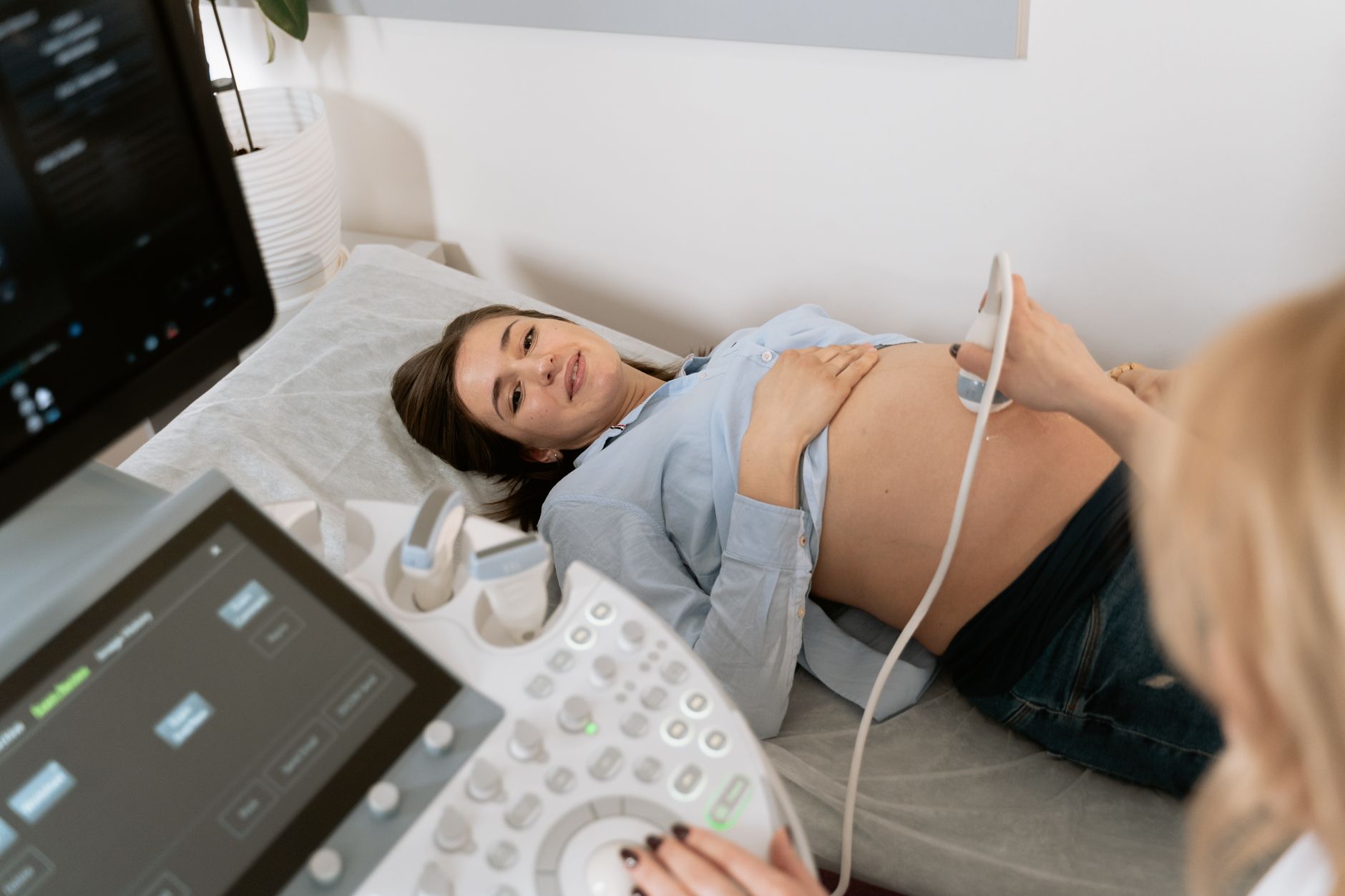 An expectant mother lays on a bed, smiling during an ultrasound in a well-lit clinic.