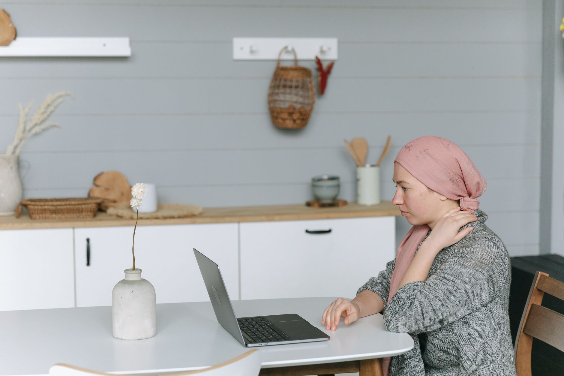 A woman with a pink scarf sits at a table, using a laptop for a follow-up video call in a cozy home setting.