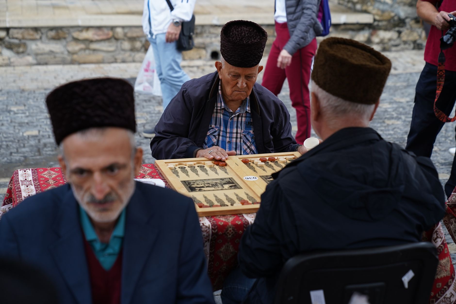 İki yaşlı adam, geleneksel şapkalar takarak bir meydandaki küçük masada tavla oynuyor.