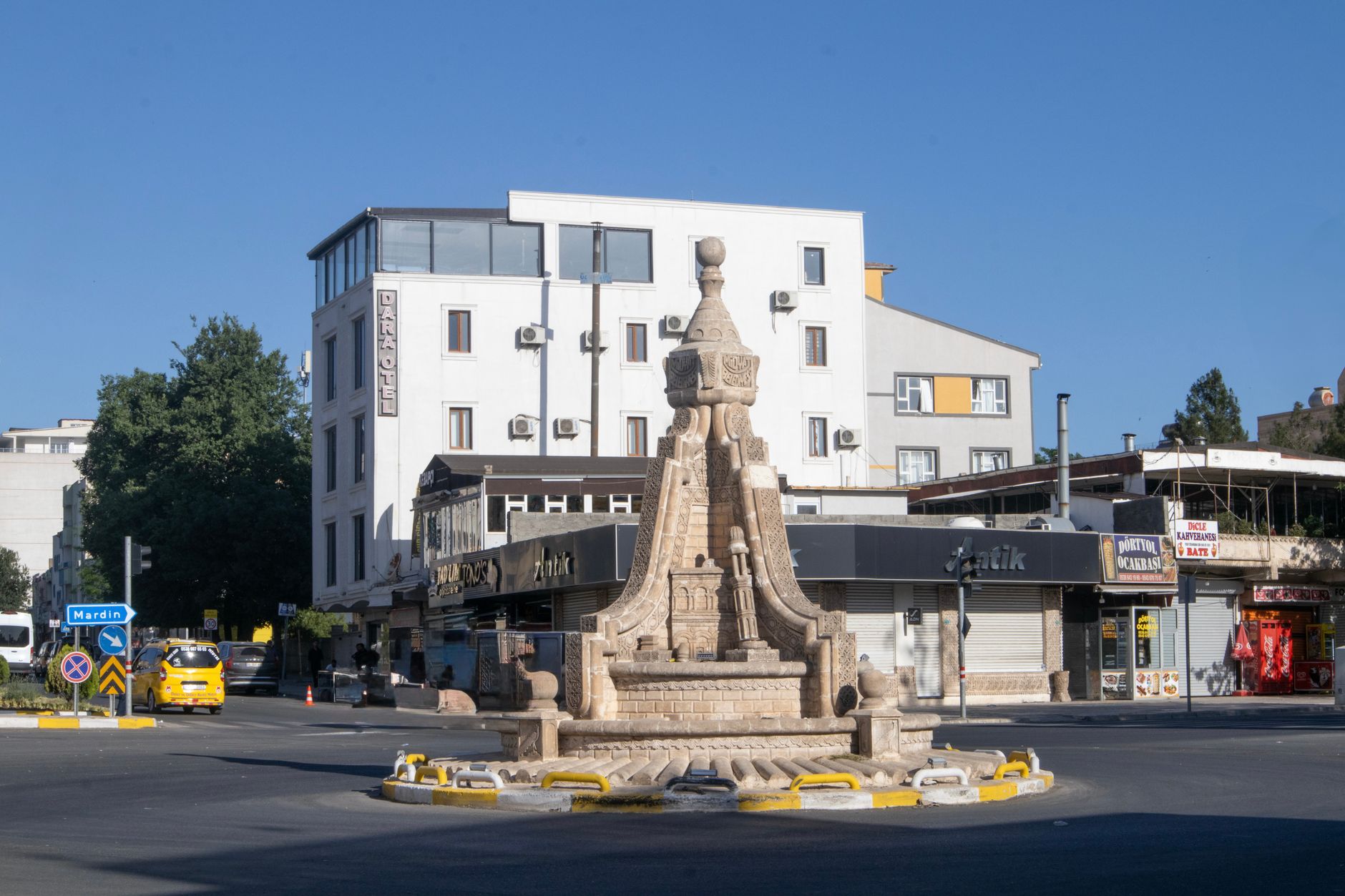 Mardin, Türkiye'de güneşli bir kasaba meydanı; dekoratif bir çeşme ve çevredeki binalar.
