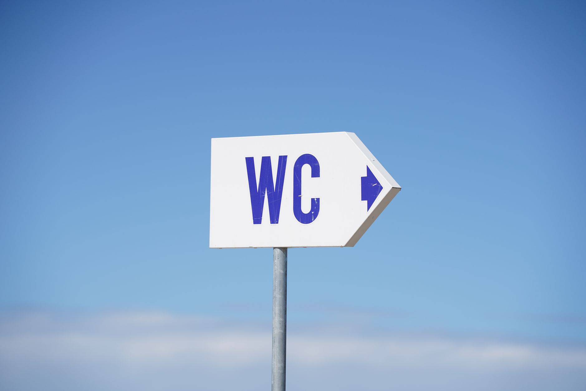 تابلوی سفیدی با نوشته «WC» و فلشی که به راست اشاره می‌کند، زیر آسمان آبی صاف.
