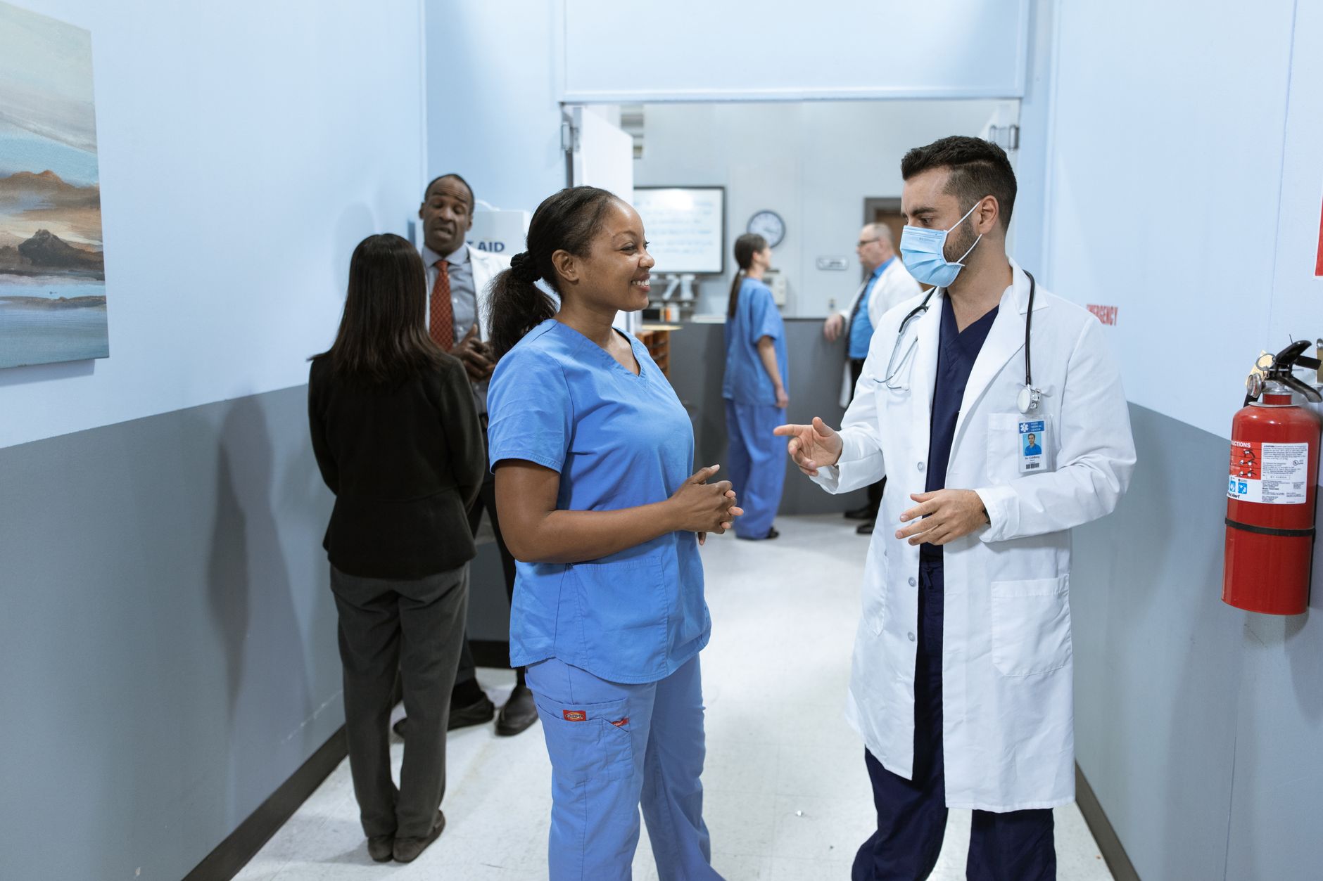 A doctor and nurse converse in a hospital hallway, with staff in scrubs and a fire extinguisher visible.