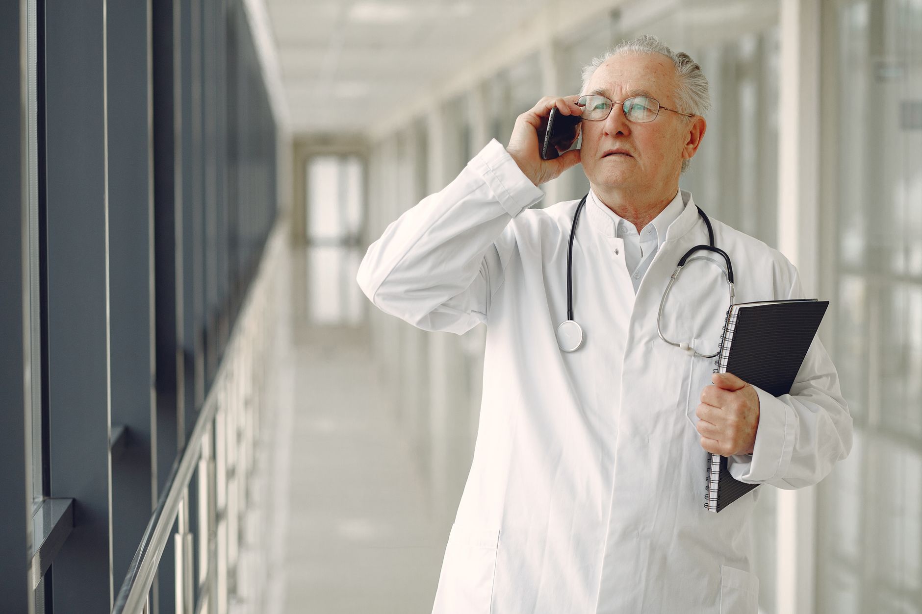 A surgeon in a white coat, holding a notebook and phone, stands in a hospital corridor.