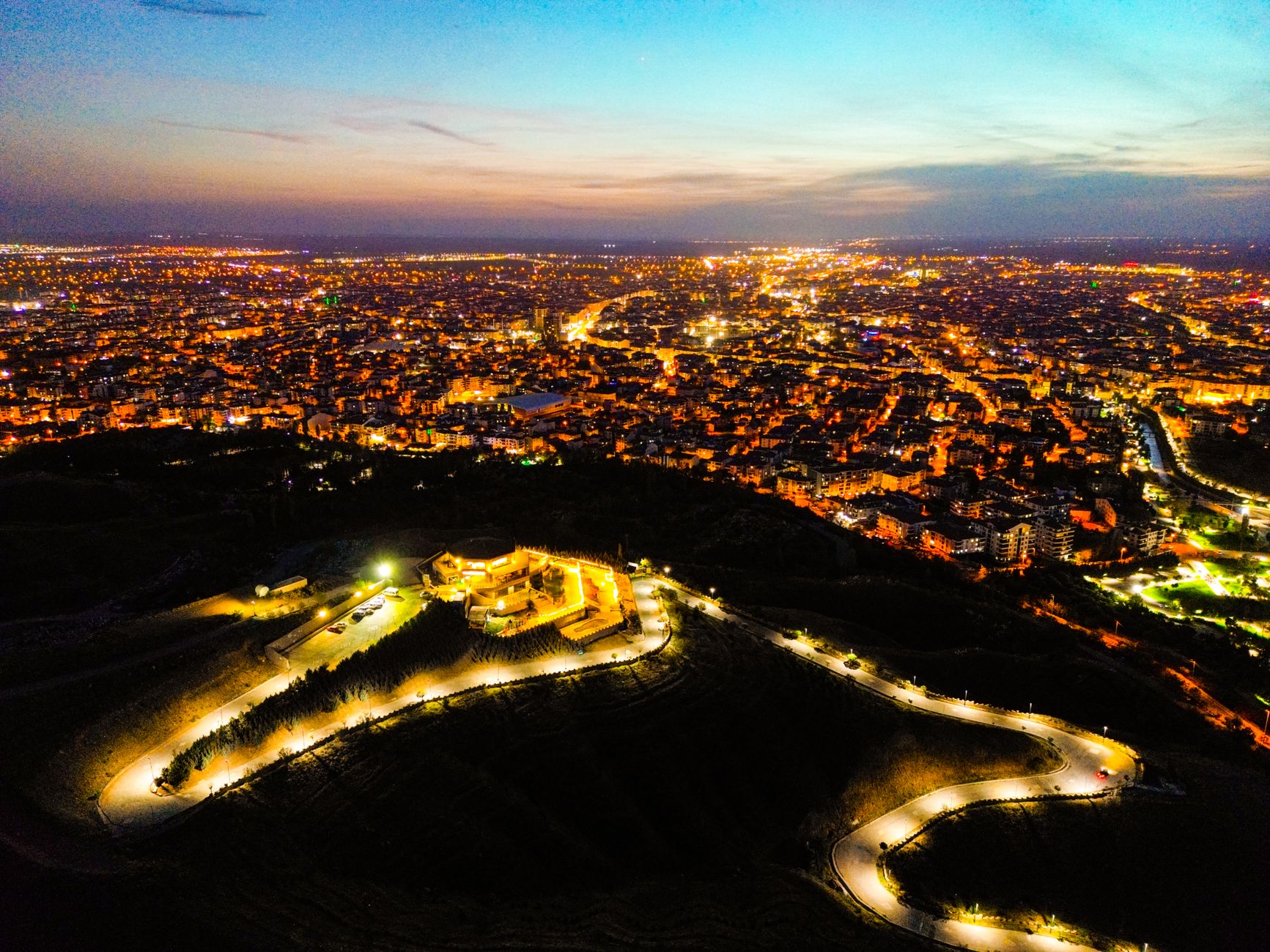 Aerial view of Gaziantep at dusk, showcasing illuminated buildings and winding roads.