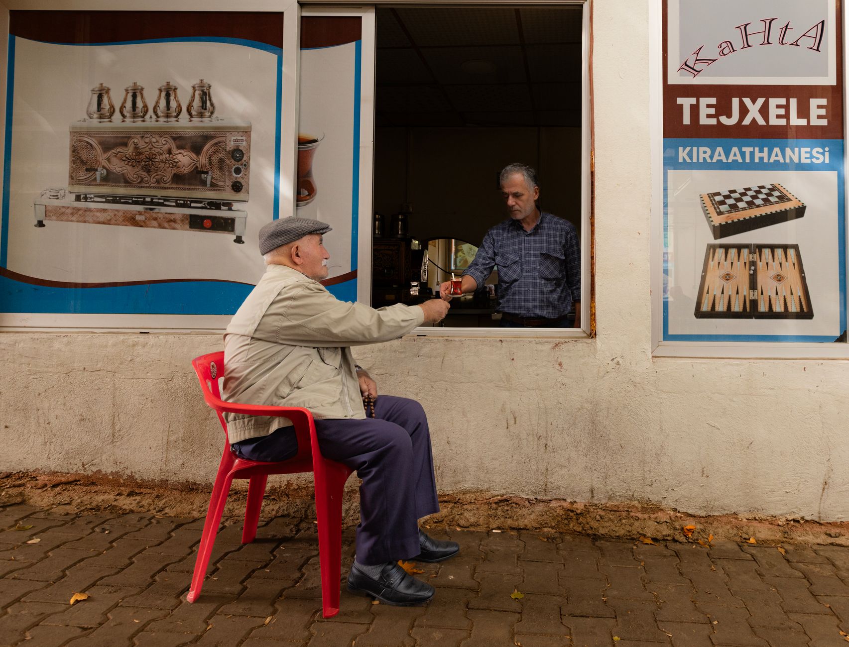 An elderly man sits in a red chair, enjoying tea while interacting with a vendor at a café.