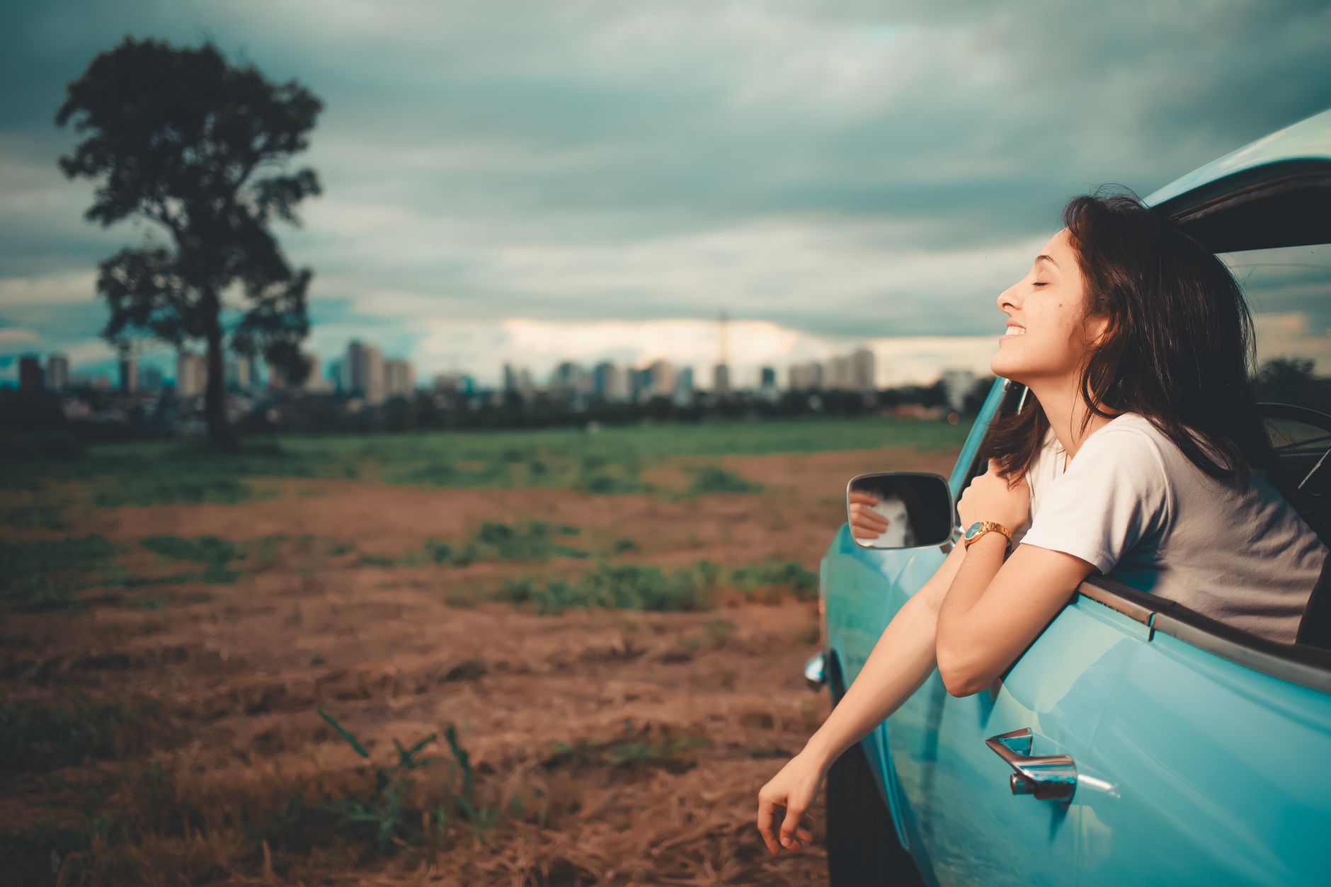 A woman relaxes with her head out of a blue car window in a field, symbolizing fresh air and improved life quality.