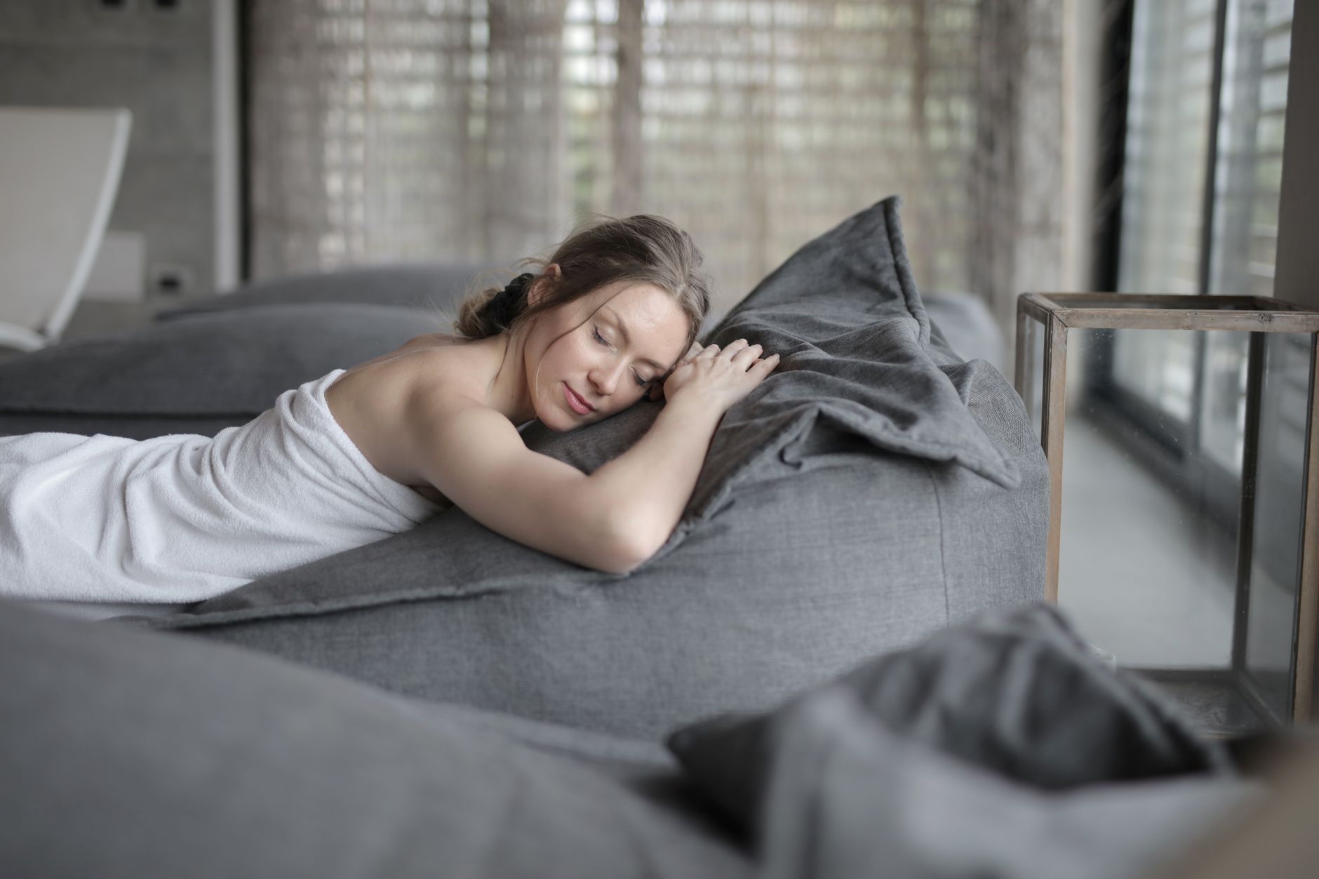 A woman wrapped in a towel peacefully rests on gray cushions in a spa-like room.