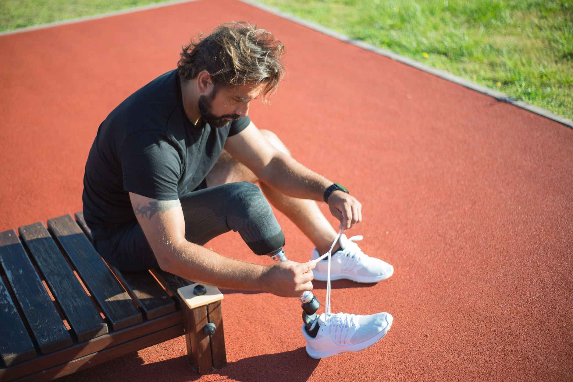 Athlete with a prosthetic leg sitting on a bench tying shoes on a red track.