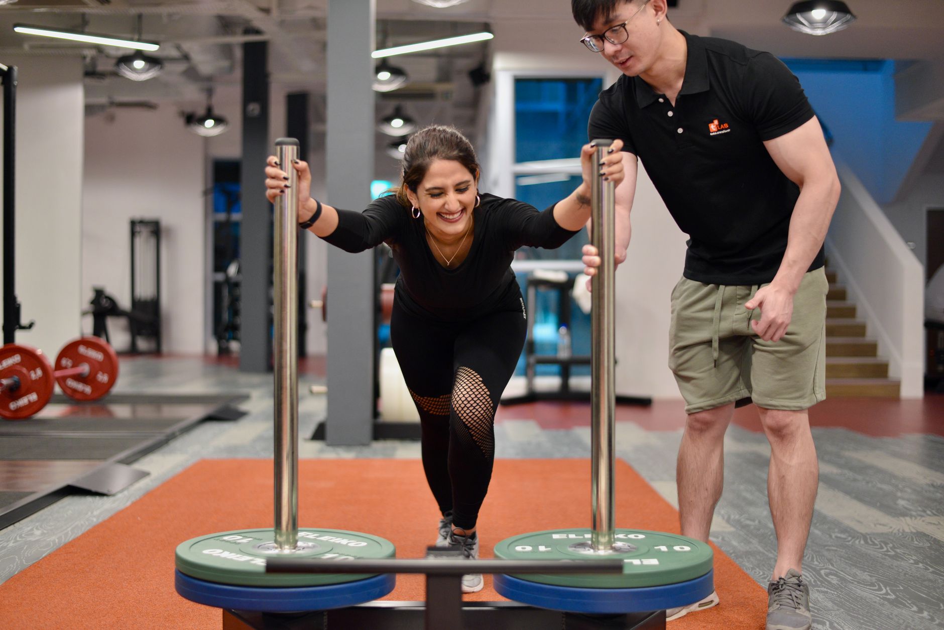 A performance coach assists an athlete in a gym during a strength training session, focusing on sled pushing.