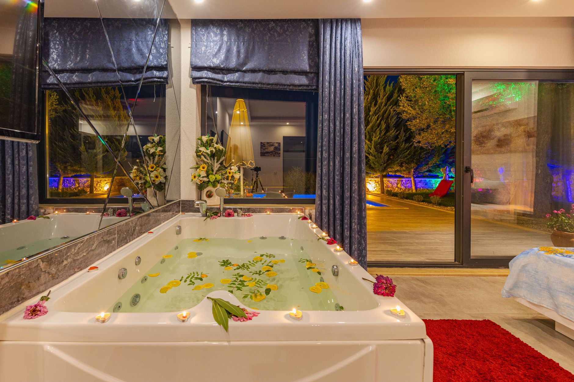 A luxurious spa bath adorned with flowers and candles, overlooking a nighttime view of a pool and garden.