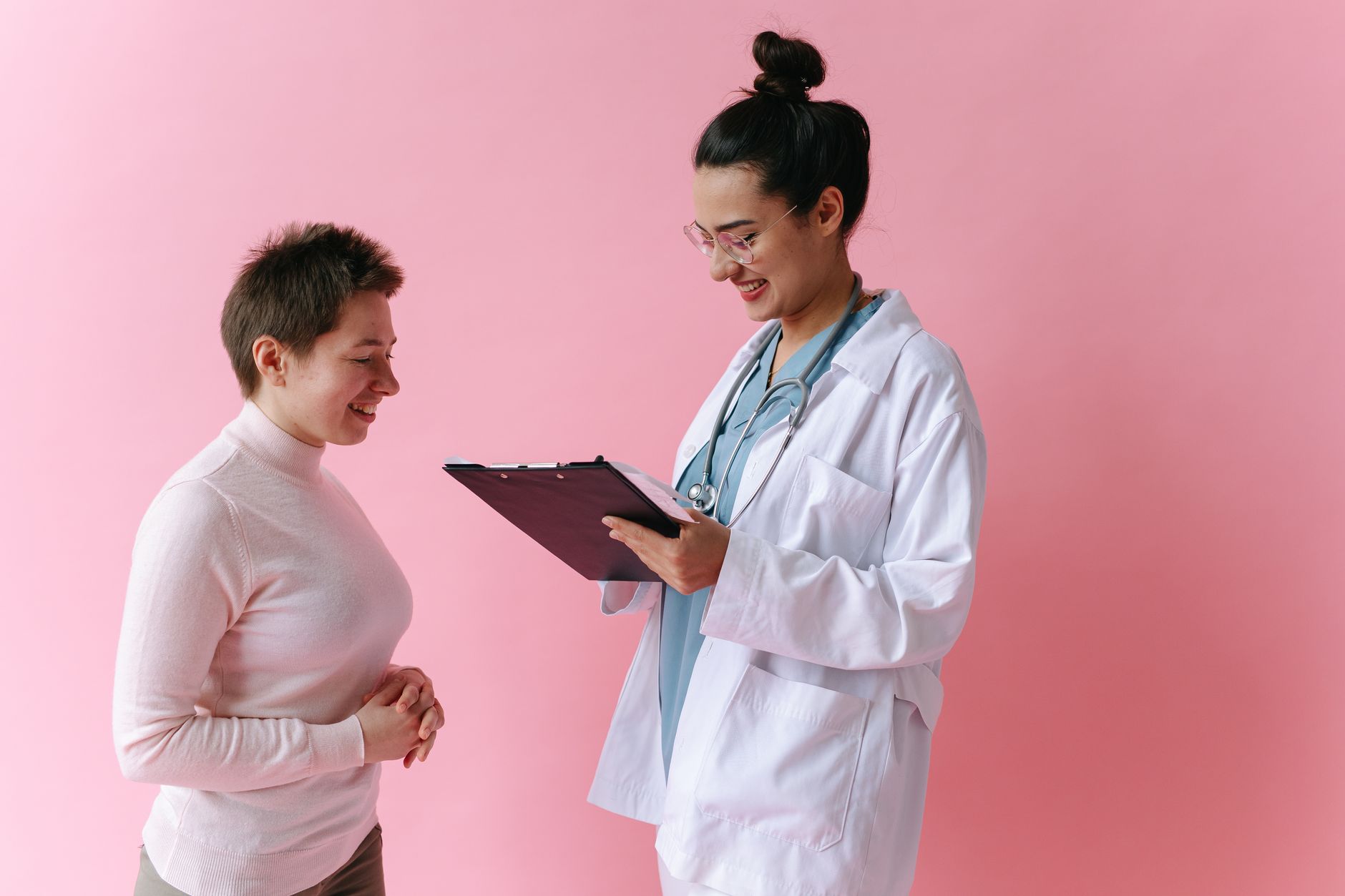 A Turkish doctor in a white coat smiles while consulting an international patient in a pink backdrop.