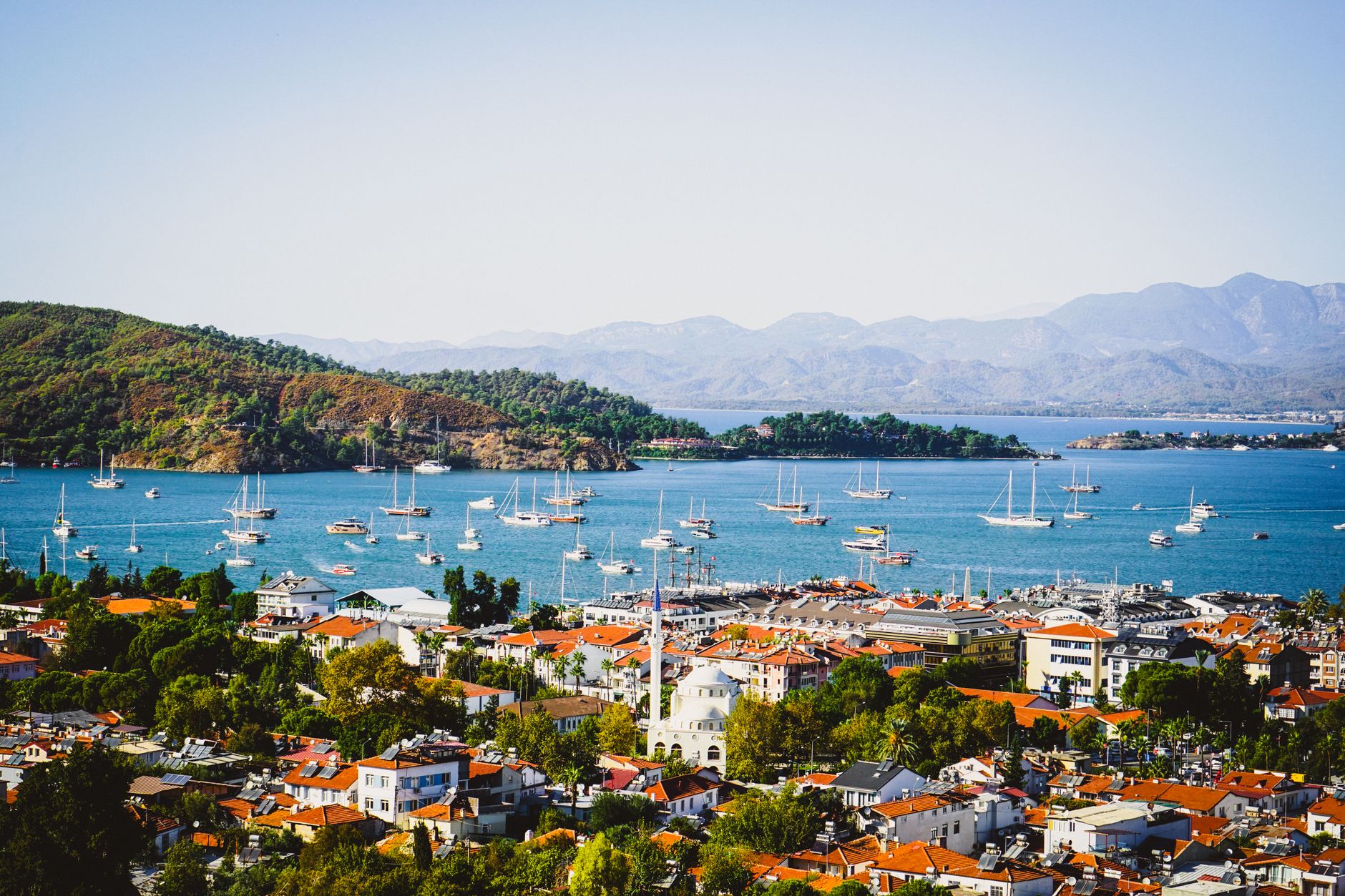 A picturesque view of the Turquoise Coast with sailboats in the blue bay, surrounded by mountains and scenic town rooftops.