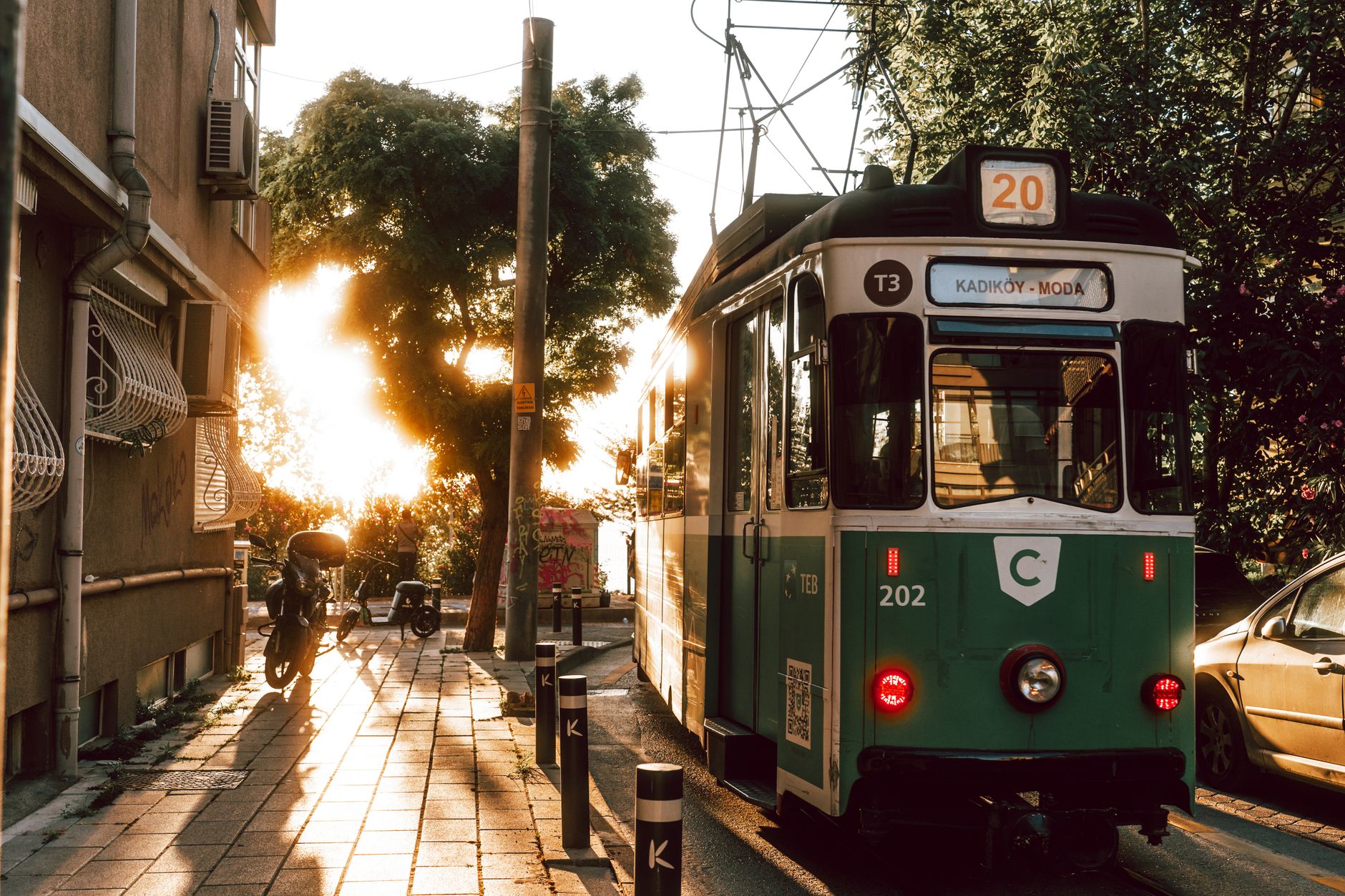 T3 tram in Kadıköy, sun setting through trees, lively street scene with shadows and nearby parked bikes.
