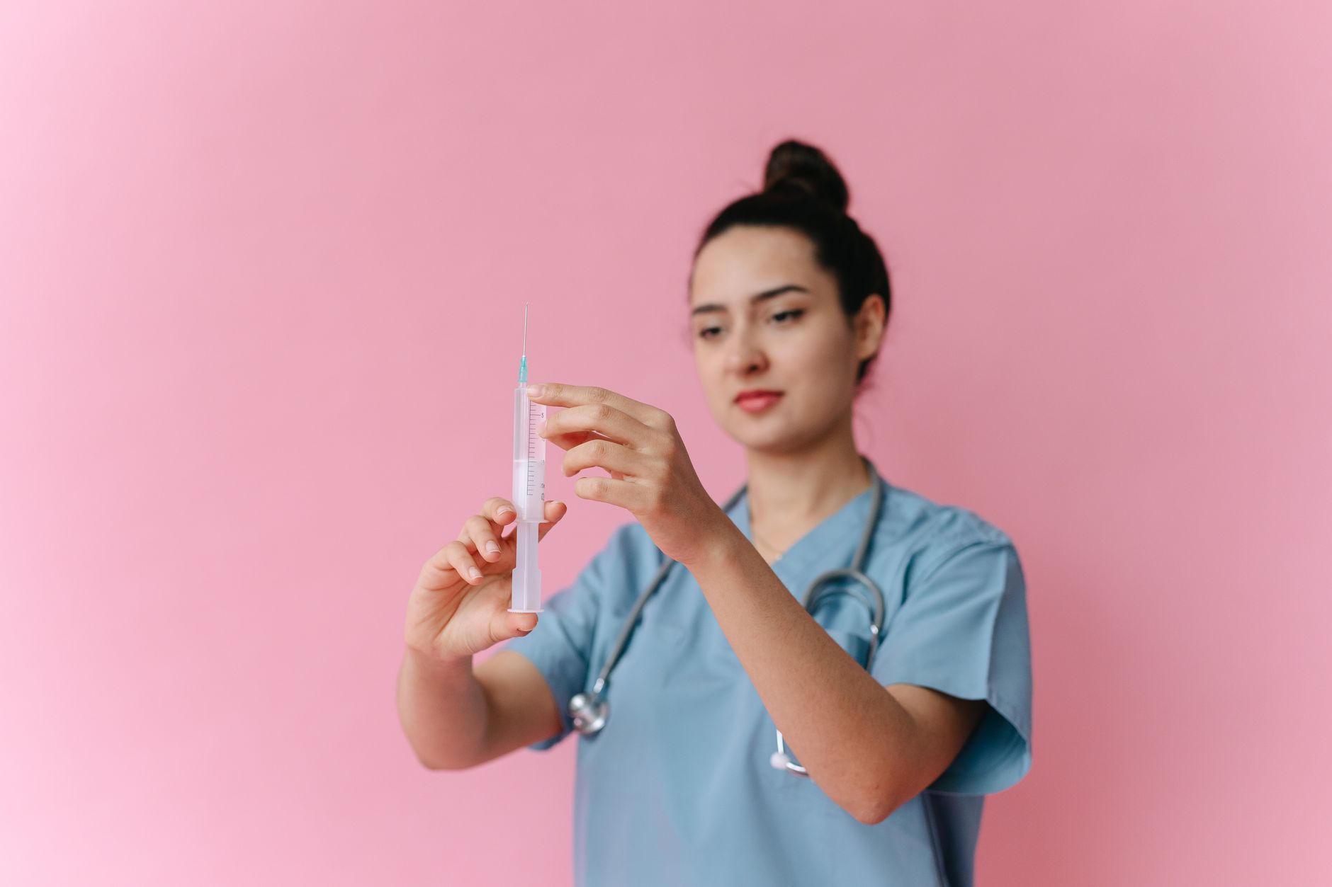A healthcare worker holds a syringe in front of a pink background, preparing for a vaccination.