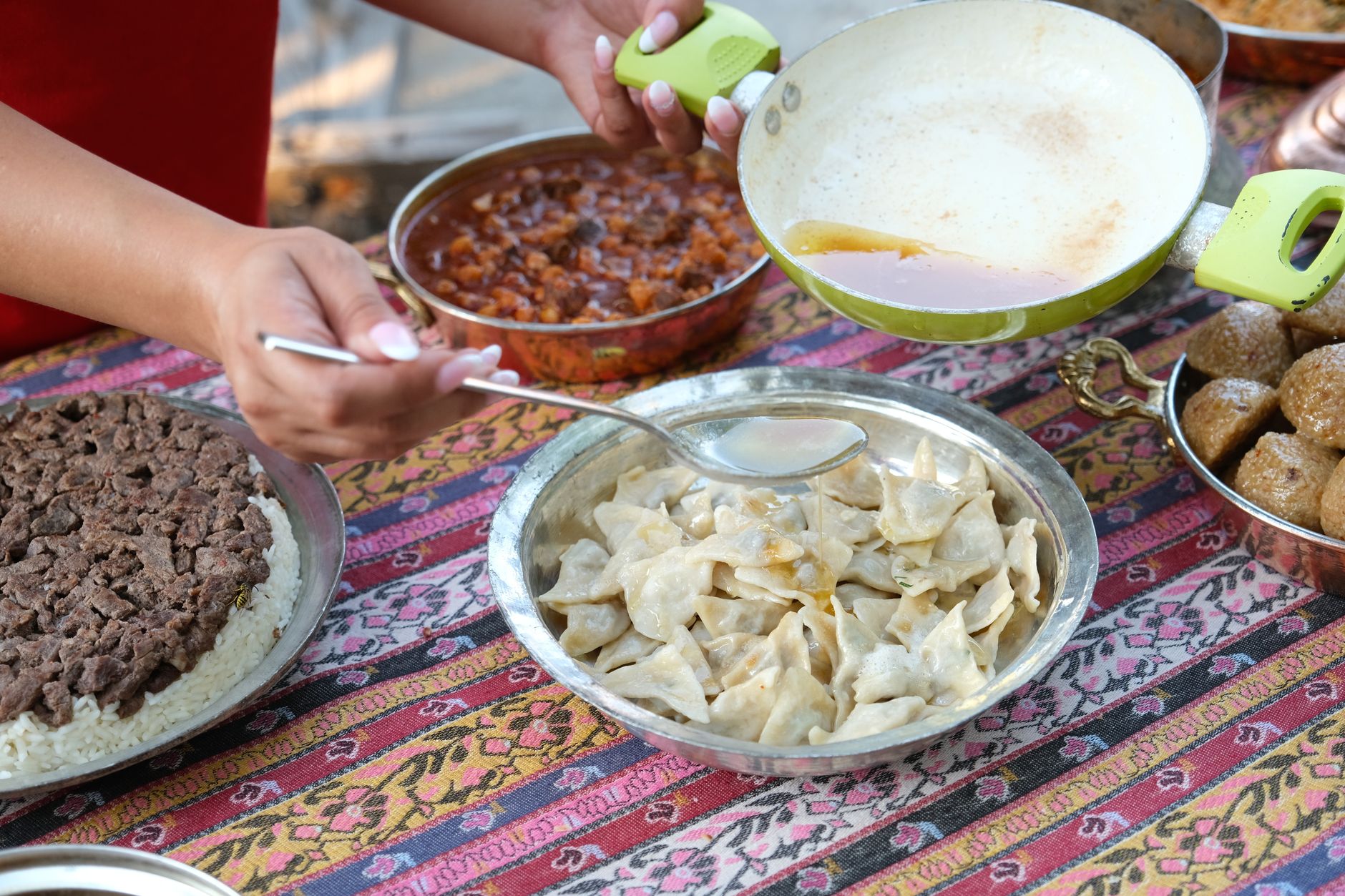 نمای نزدیک از دست‌هایی که کوفته‌های مانتی را شکل می‌دهند، همراه با غذاهای مختلف روی رومیزی نقش‌دار.