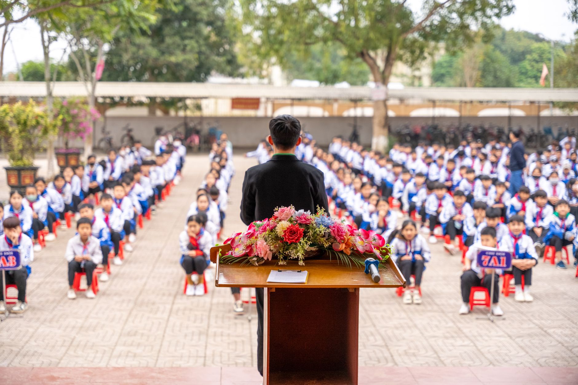 A teacher speaks at a podium in front of a large group of seated students.