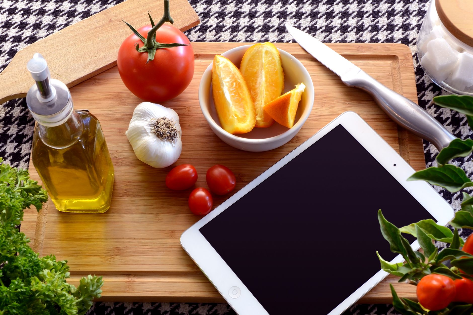 Wooden cutting board with fresh vegetables, olive oil, and a tablet for recipe guidance.