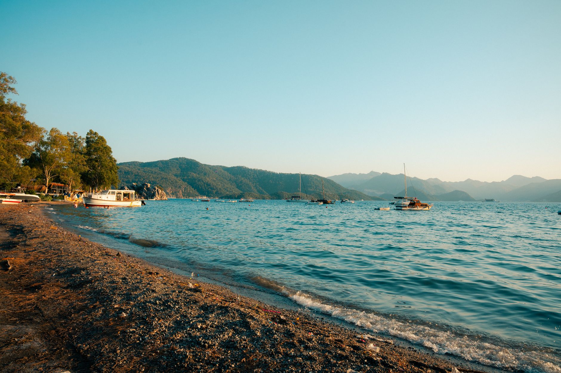 A serene beach in Marmaris with boats on turquoise Mediterranean water and distant mountains.
