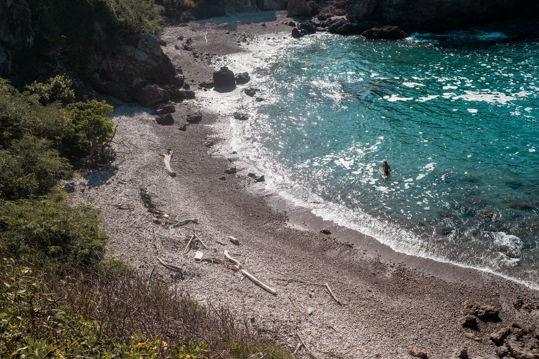 A tranquil Mediterranean cove featuring clear turquoise water, rocky shoreline, and beach debris.