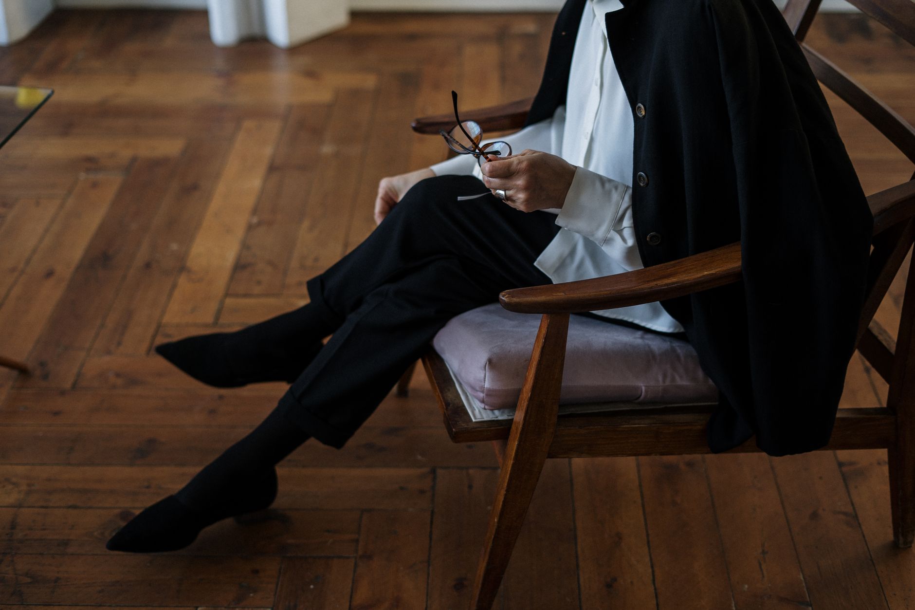 Close-up of a person seated in a wooden chair, wearing black attire and holding glasses.