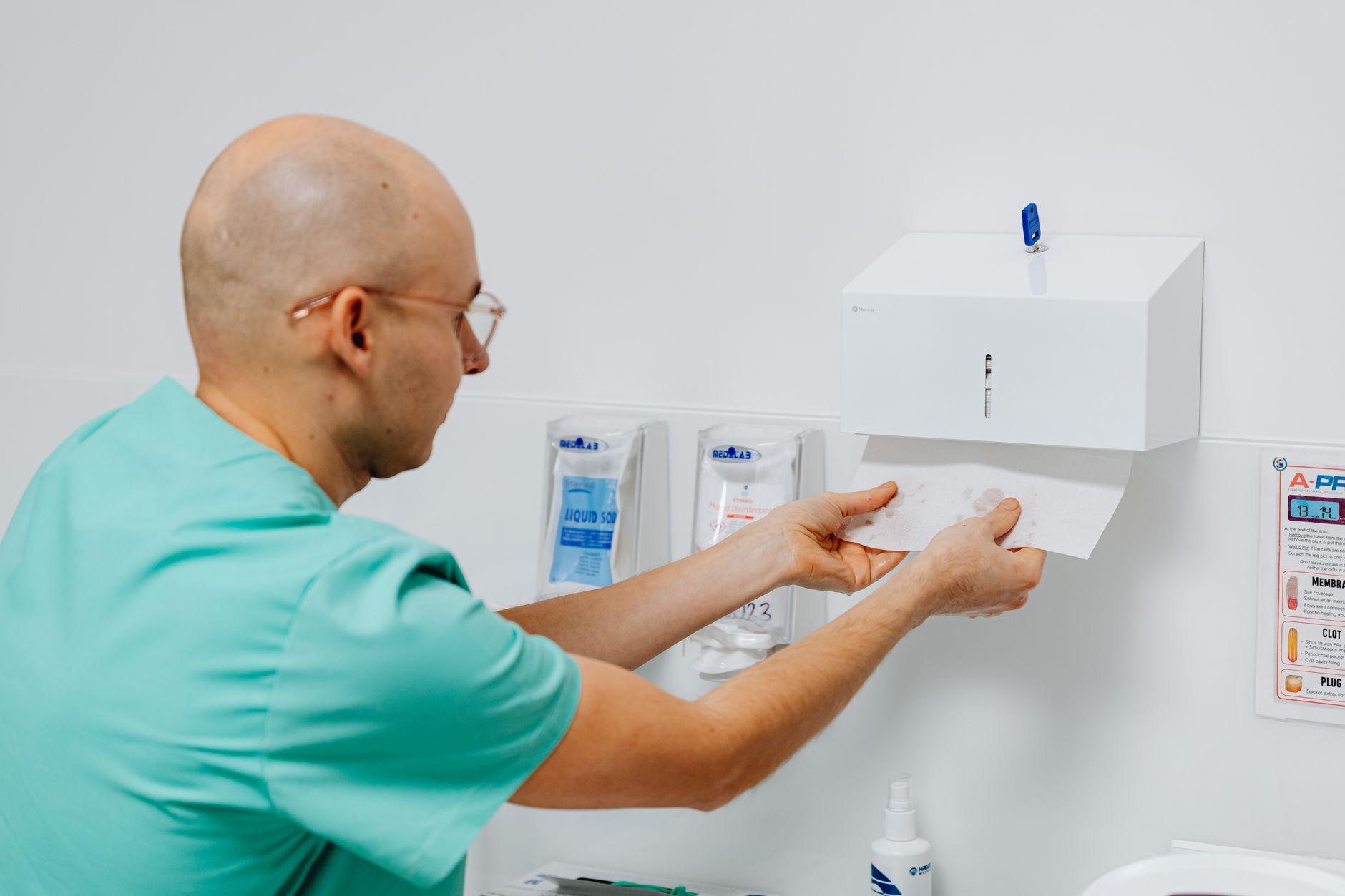 A healthcare worker in scrubs dispensing paper towels from a wall-mounted sanitizer in a hospital setting.