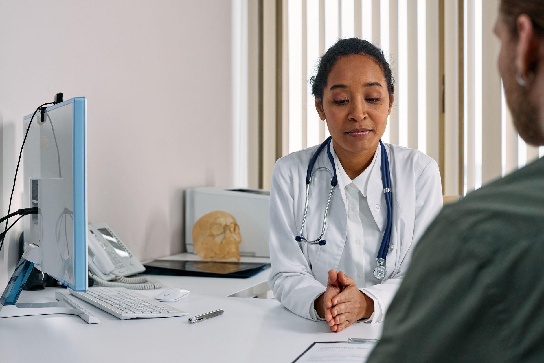 A female doctor in a white coat reviews notes with a patient in a bright clinic office.